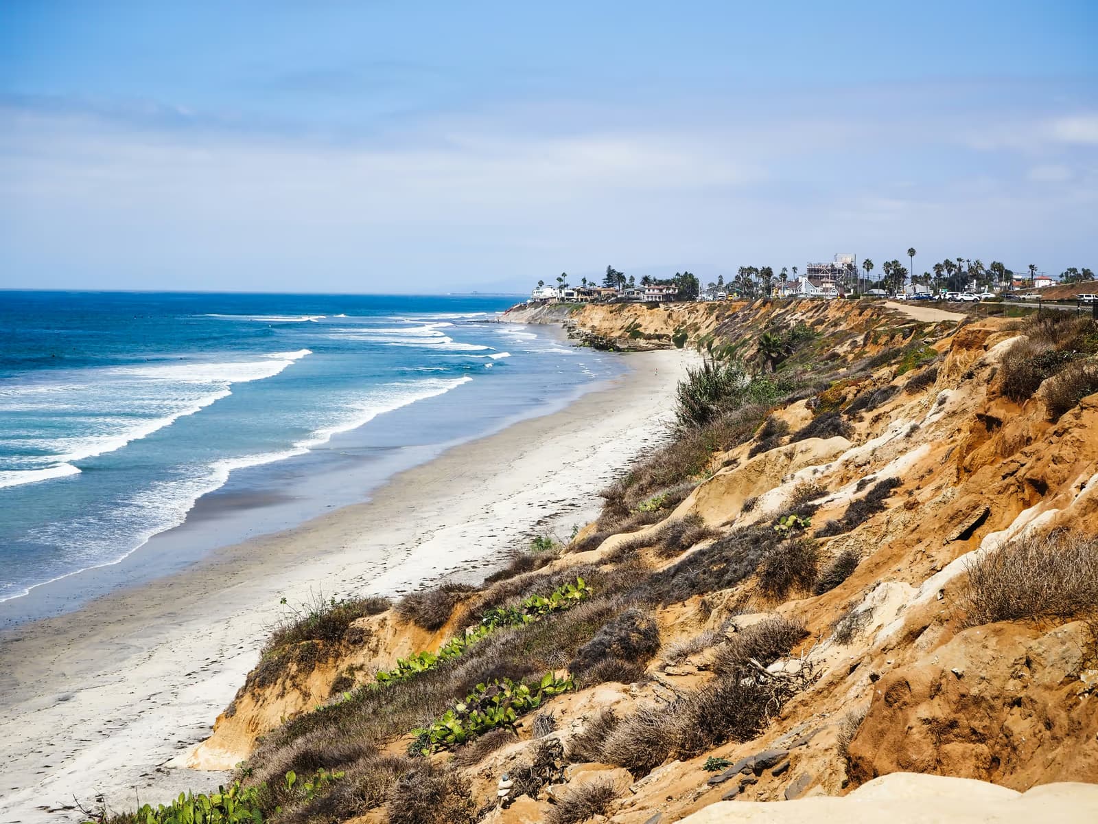 Waves breaking on a long sandy beach below eroded sandstone bluffs, with houses and palm trees along the clifftop