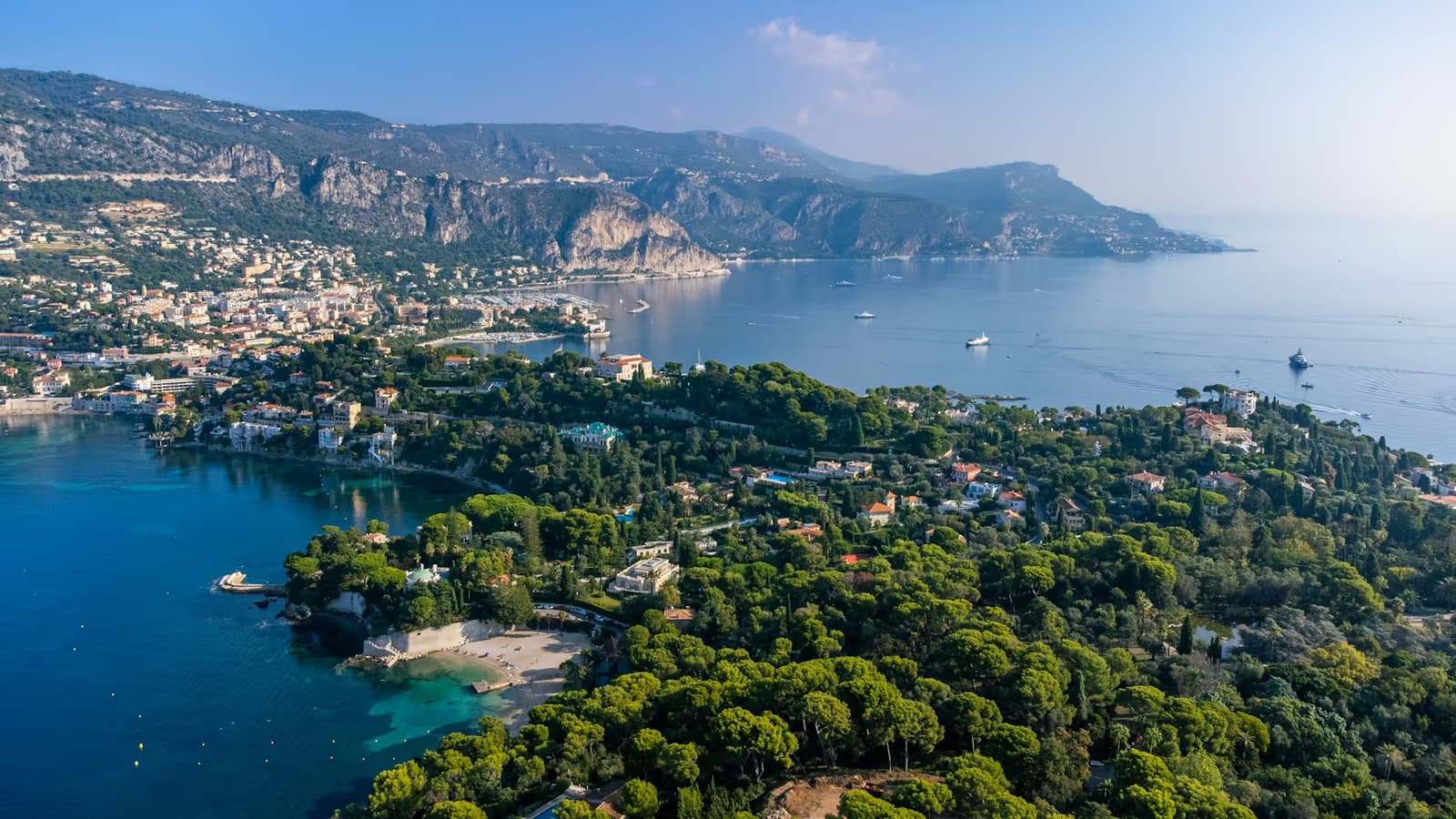 Pine woods and villas on the Saint-Jean-Cap-Ferrat peninsula above a blue bay, with the mountains of the Riviera behind