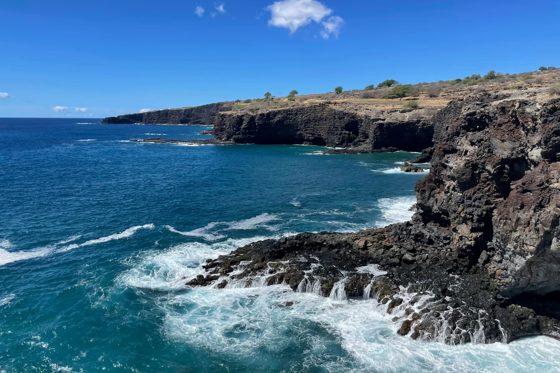 Waves breaking against dark lava sea cliffs under a blue sky on the coast of Lanai