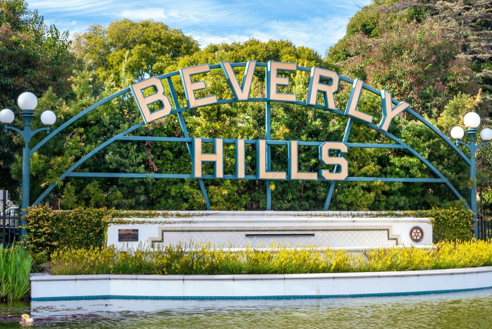 The arched Beverly Hills sign above a lily pond, with trees and yellow flowers behind