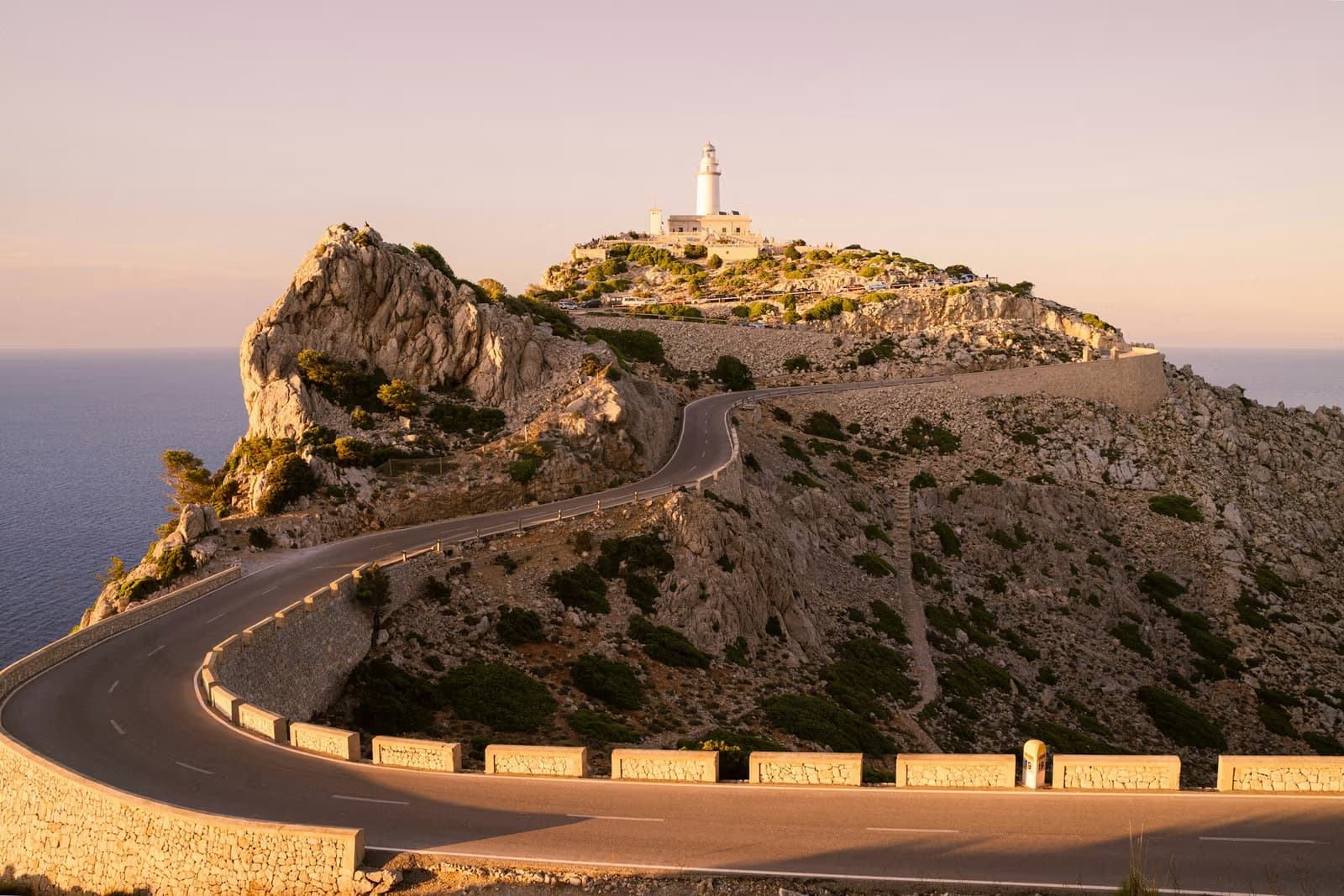 A walled road curving along a rocky headland to a white lighthouse above the sea at sunset