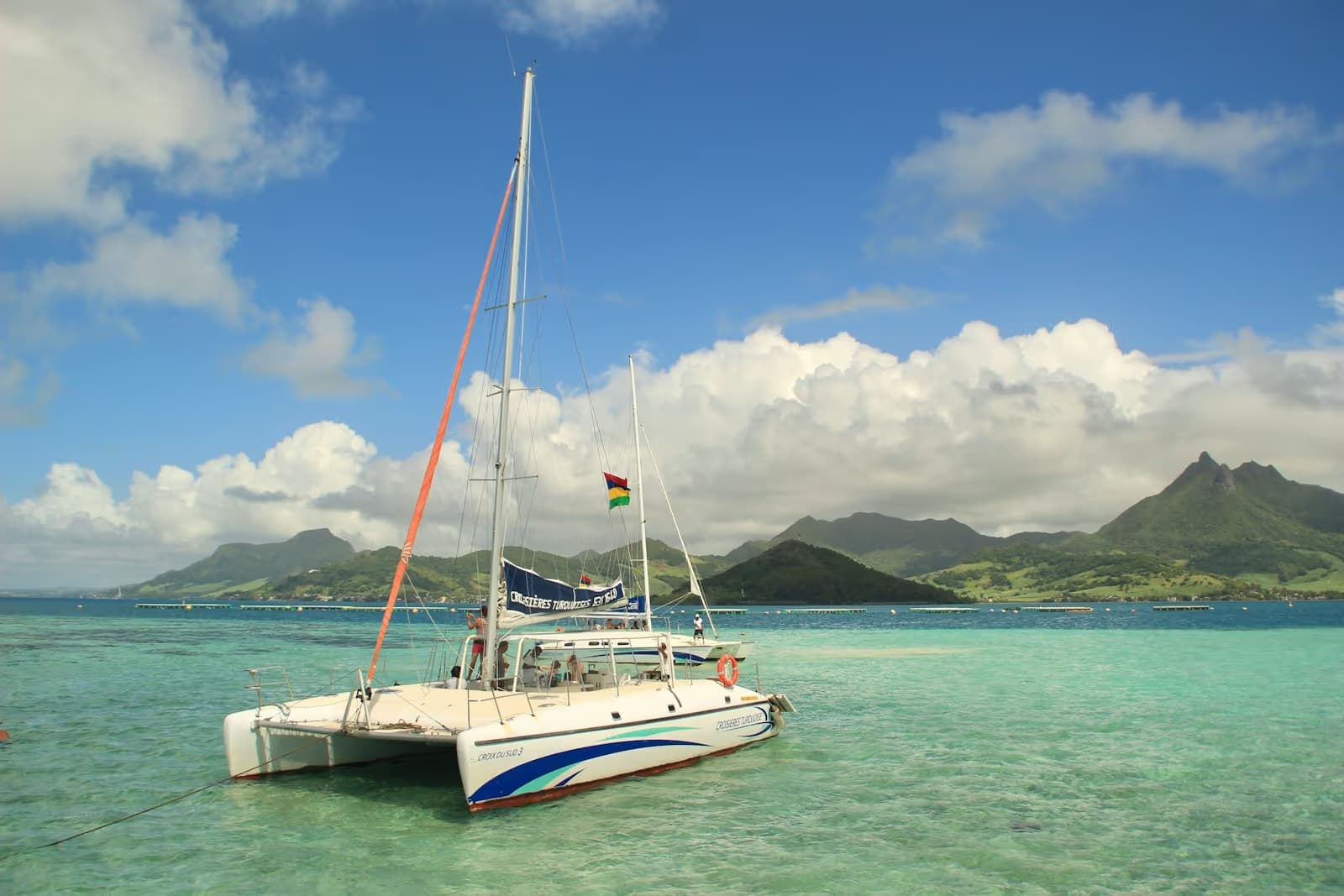 A catamaran anchored in a shallow turquoise lagoon, with green mountains behind