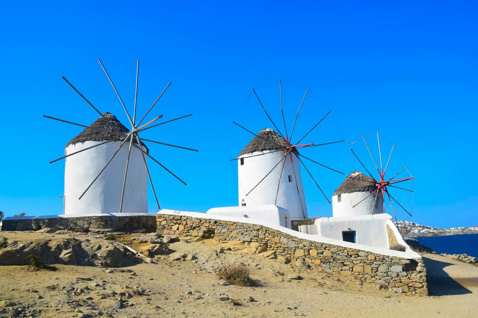 Three white windmills with thatched roofs on a stone terrace above the sea, under a clear blue sky