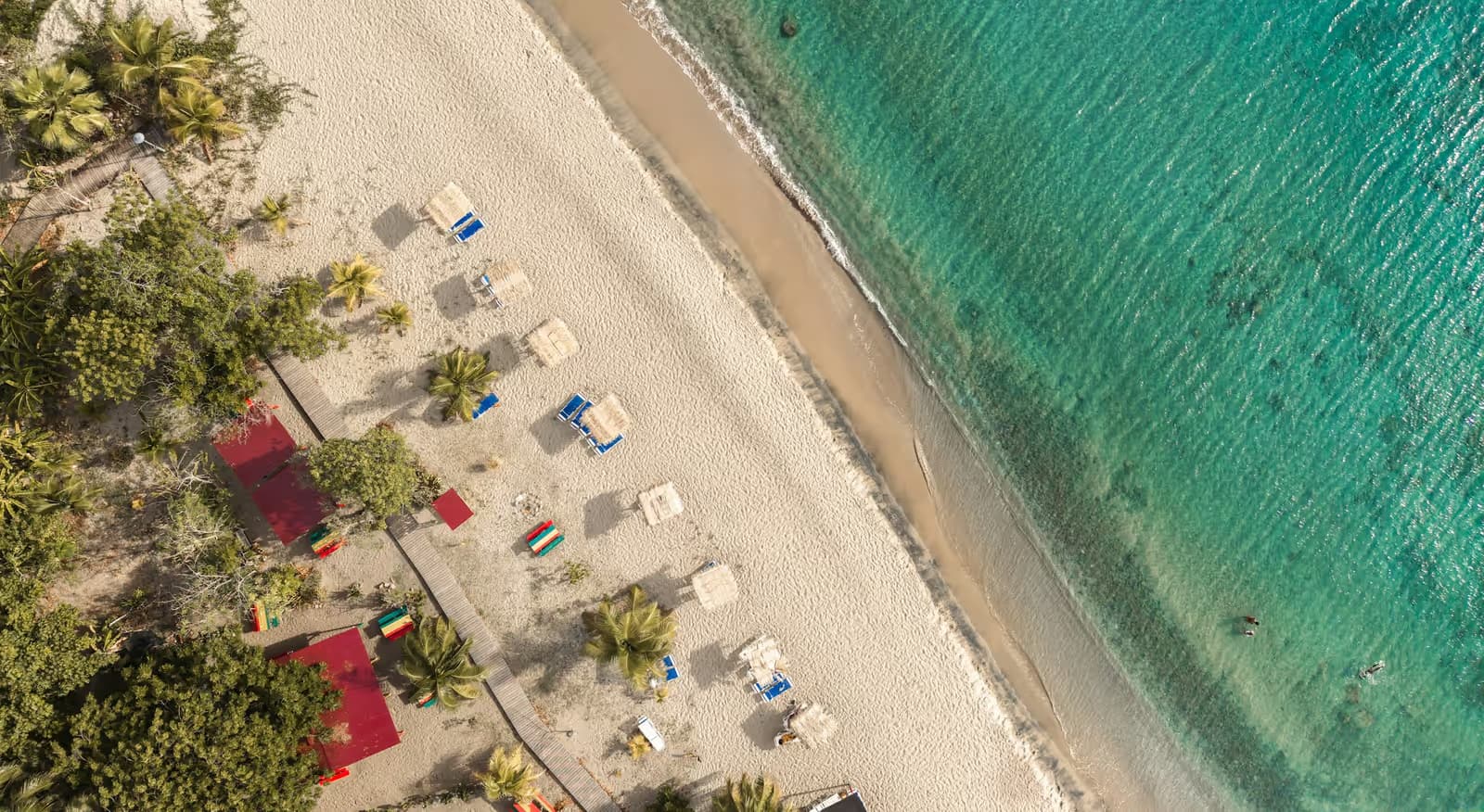 Aerial view of a white-sand beach with palm trees, loungers and red-roofed beach bars beside turquoise water