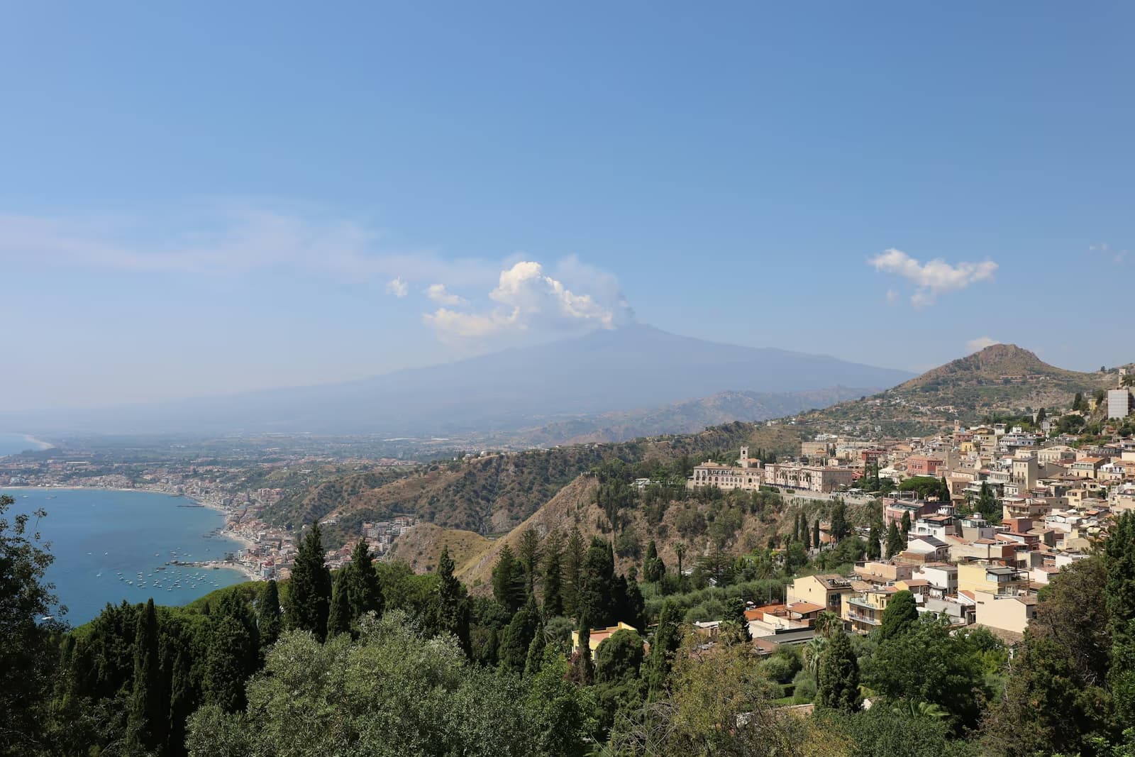 Taormina's hillside houses above the coast, with Mount Etna and a plume of cloud in the distance