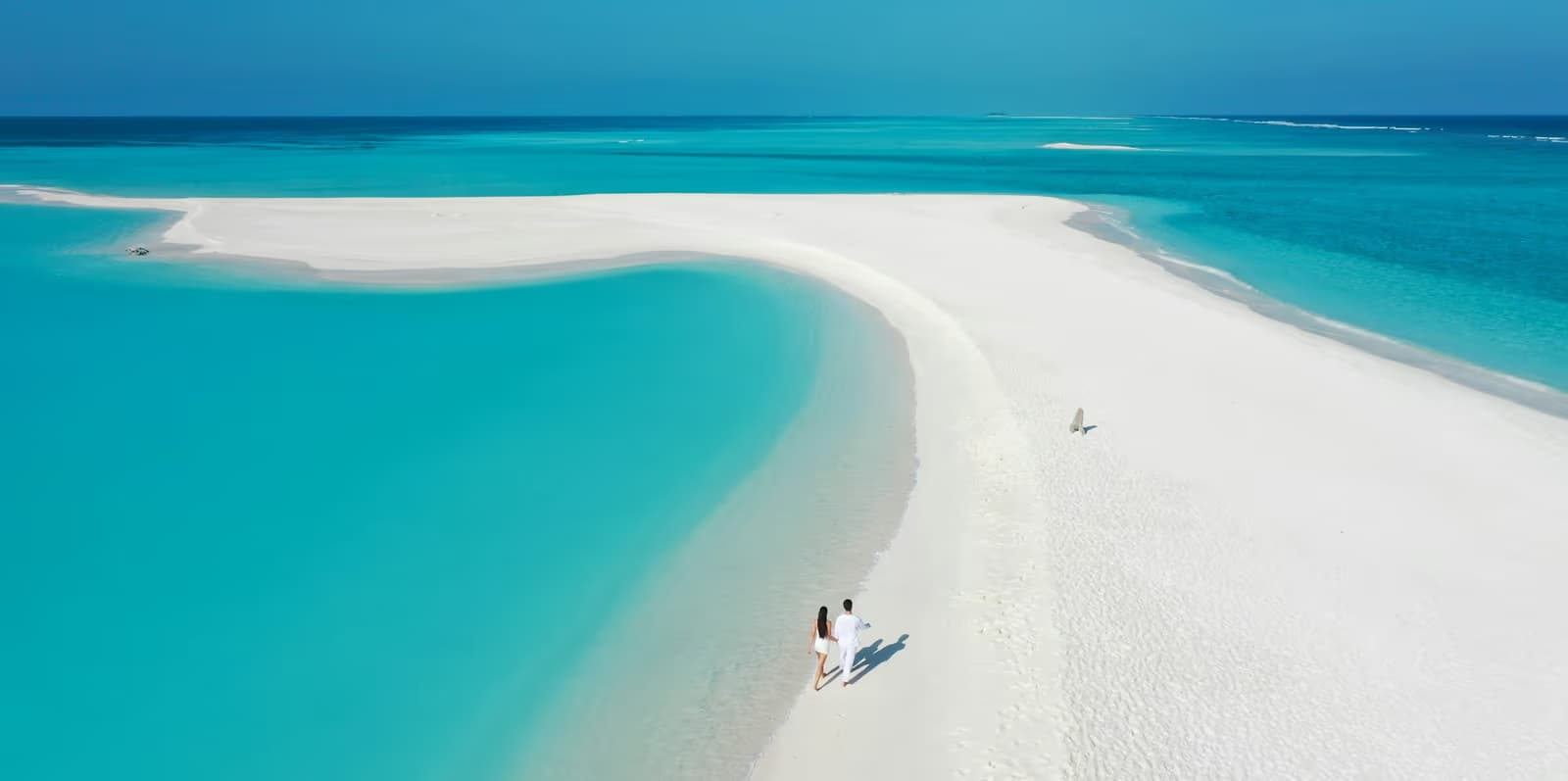 Aerial view of two people walking along a long white sandbar between turquoise lagoon and open sea