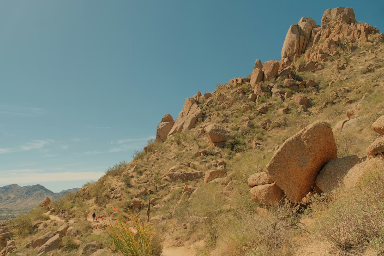 A hiking trail climbing a desert hillside of granite boulders and dry scrub under a clear blue sky, with mountains in the distance