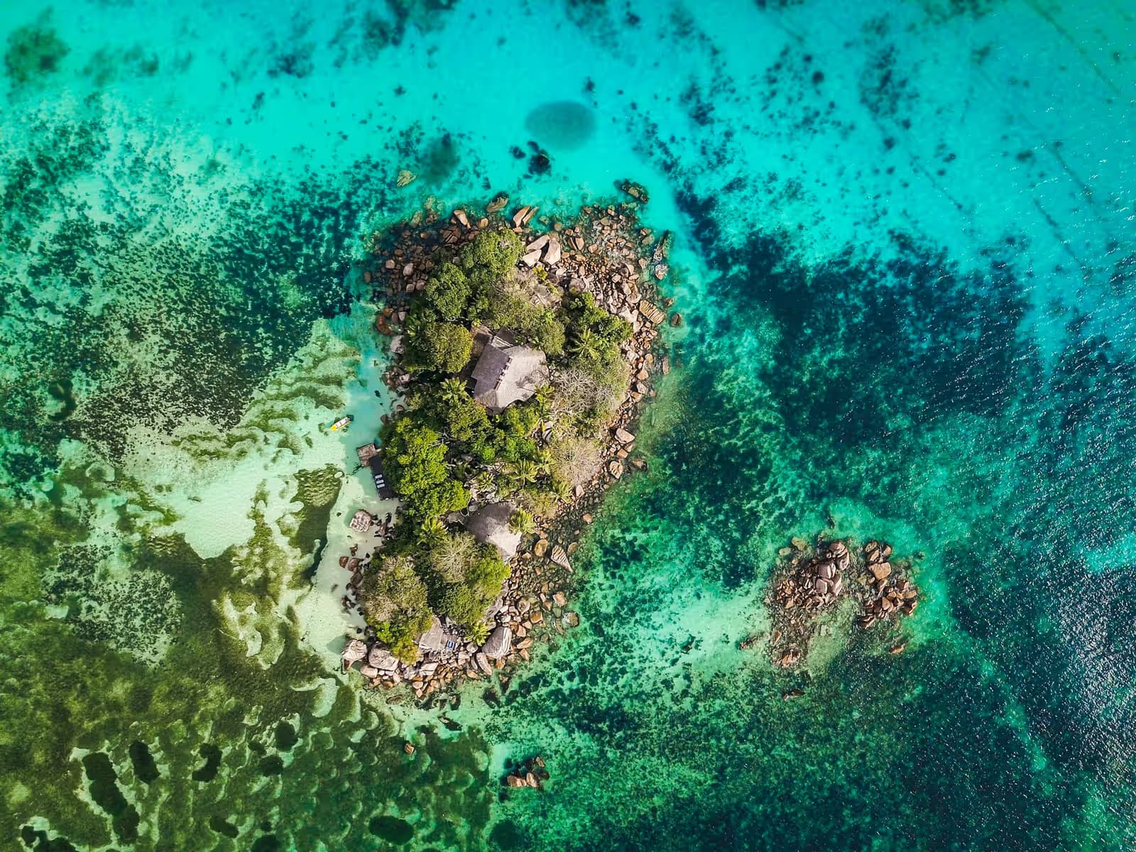 Aerial view of a small rocky islet with palms and a few buildings, surrounded by turquoise reef water