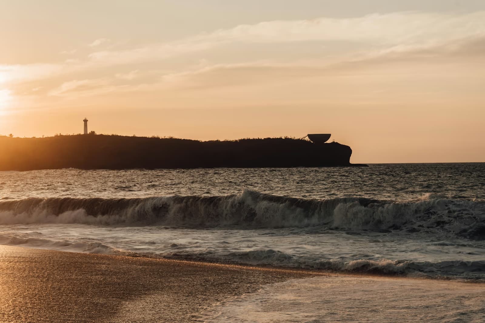 Waves breaking on a sandy beach at sunset, with a dark headland topped by a lighthouse and a bowl-shaped sculpture